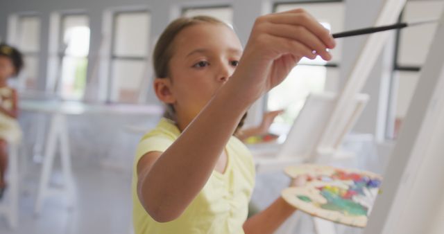 Young girl painting on canvas in an art classroom, holding a palette and paintbrush. Ideal for use in educational materials, creative learning content, art class promotions, and teaching resources that emphasize creativity and artistic development in children.