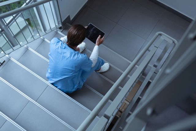 A healthcare professional in surgical scrubs is using a digital tablet while sitting on stairs in a hospital. This image can be used in articles or advertisements related to medical technology, healthcare environments, or the importance of breaks for healthcare workers. Ideal for depicting modern medical practices and the integration of technology in healthcare.