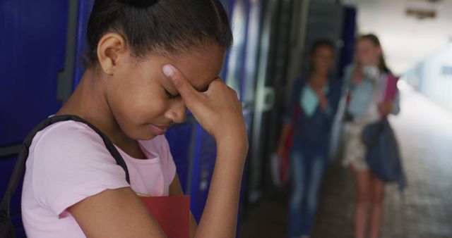 Young student is standing near lockers in a school hallway, appearing worried and stressed. She is holding books and touching her forehead, possibly indicating distress or anxiety. Other students can be seen walking in the background, providing a school environment context. This image can be used to illustrate concepts related to school stress, bullying, teenage emotions, and mental health awareness.