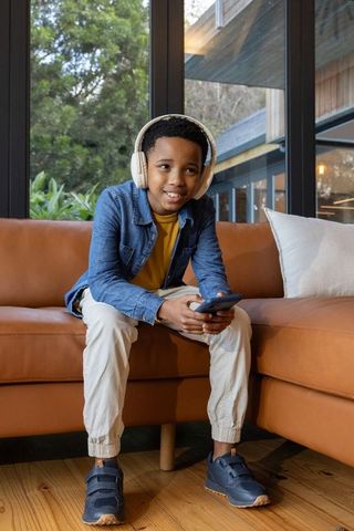 African american preteen boy sitting on sofa listening with headphones holding smartphone