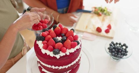 Friends decorating red velvet layer cake with fresh berries on bright kitchen countertop