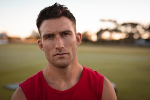 This image features a fit Caucasian man wearing a red tank top, looking directly at the camera with a serious expression. The background shows an outdoor sports field, suggesting a fitness or training environment. This image can be used for promoting fitness programs, athletic apparel, health and wellness campaigns, or motivational content related to exercise and physical training.