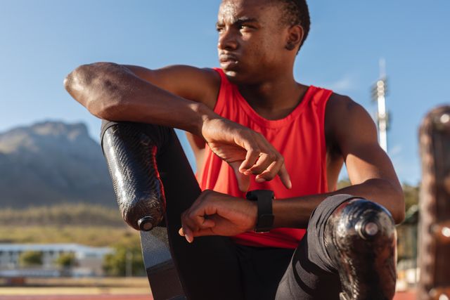 This image shows a fit and focused biracial disabled male athlete sitting on a race track at an outdoor sports stadium, wearing running blades. Ideal for use in articles or advertisements about disability sports, athletic training, motivation, and overcoming challenges. Suitable for promoting sportswear, fitness equipment, and inspirational stories.