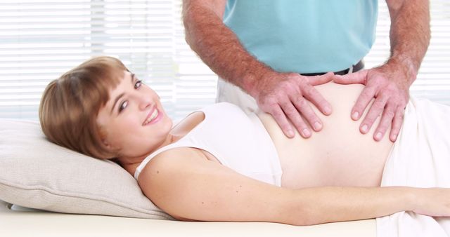 Male caucasian therapist touching belly of smiling caucasian pregnant woman with copy space. Motherhood, pregnancy, therapy and wellness concept, unaltered.