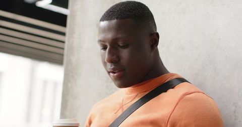 African American man leaning against concrete wall holding coffee in orange sweatshirt