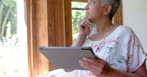 Pensive Senior Woman Using Tablet by Window at Home