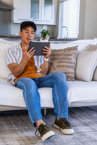 Asian man in 30s relaxing on sofa using tablet in modern bright living room