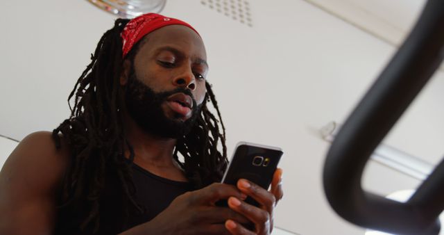 An African American man with dreadlocks is focused on his smartphone, managing his fitness routine or tracking his workout progress. His engagement with technology underscores the modern approach to personal health and fitness.