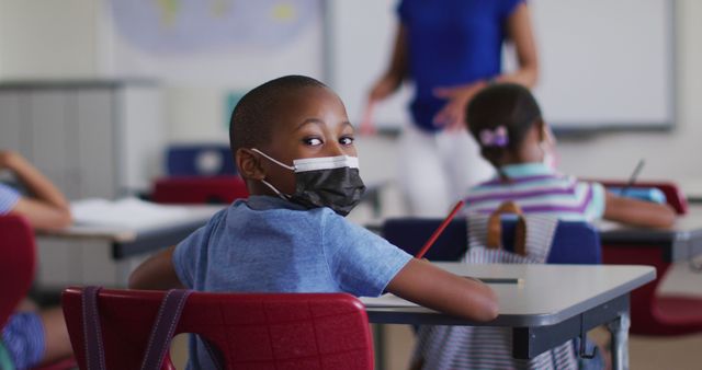 Portrait of african american schoolboy sitting in classroom, making notes wearing face mask. children in primary school during coronavirus covid 19 pandemic.