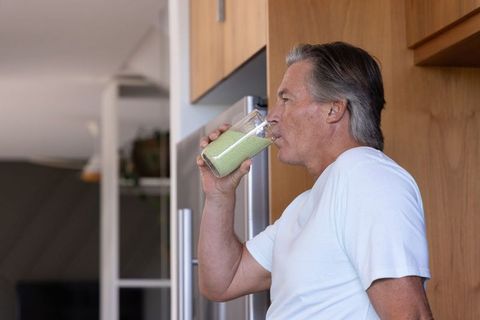 Mature man sipping green smoothie in kitchen by stainless fridge and wood cabinets