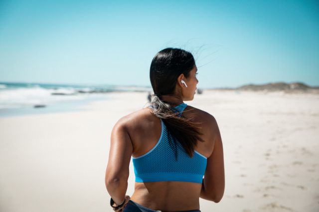 Biracial woman running on a sandy beach wearing wireless earphones, showcasing a healthy and active lifestyle. Ideal for promoting fitness, outdoor exercise, and wellbeing. Perfect for use in advertisements, fitness blogs, and health-related content.