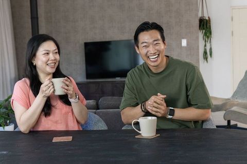 Asian couple laughing and chatting over coffee at dark wooden table in cozy living room