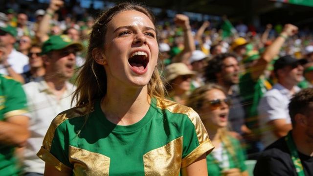 Young woman cheering energetically in green and gold fan jersey at packed outdoor stadium
