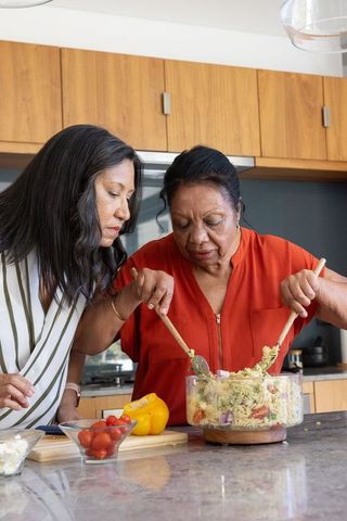 Senior mother and adult daughter mixing pasta salad during family cooking at kitchen island