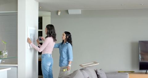 Female friends hanging picture with spirit level indoors creative collaboration