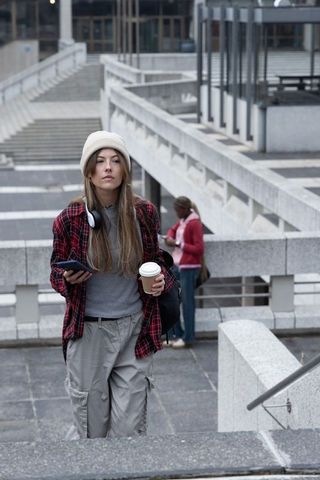 Young woman walking up concrete stairs holding coffee and smartphone wearing knit beanie