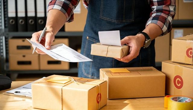Small Business Owner Packing Orders and Checking Shipping Labels in Busy Fulfillment Workspace