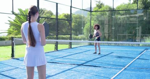 Diverse women playing padel on blue glass court during sunny outdoor workout session