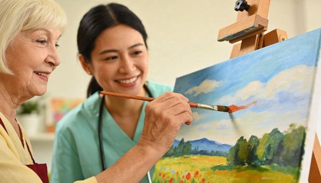 Senior woman painting landscape with nurse guiding brush during therapeutic art session
