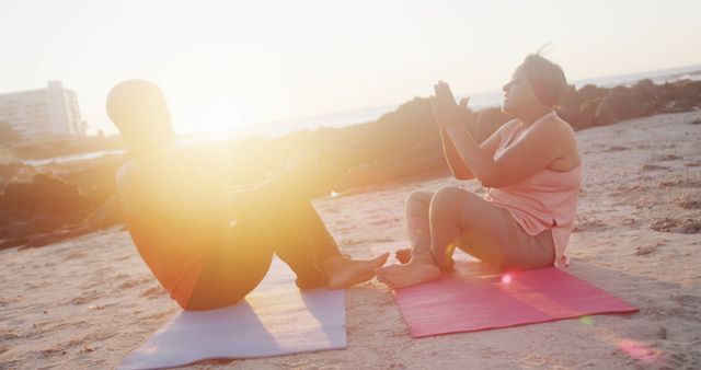 Couple practicing yoga on beach at sunset, sitting on mats, focusing on meditation and fitness. Warm sunlight creates a tranquil atmosphere, perfect for promoting relaxation and wellness. Ideal for use in fitness, wellness, and relaxation content or articles about outdoor exercises and healthy lifestyle.