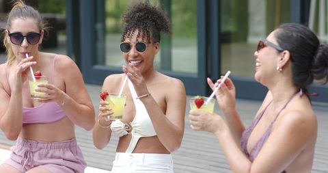 Multiethnic friends laughing and sipping cocktails on sunlit terrace in summer swimwear
