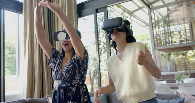 Two women wearing VR headsets are enjoying an immersive virtual reality experience in a stylish and modern home. They appear to be engaged and entertained, showcasing the joy and fun associated with VR technology. This could be ideal for promotions related to virtual reality products, tech reviews, lifestyle blogs, and advertisements depicting technology in everyday life.