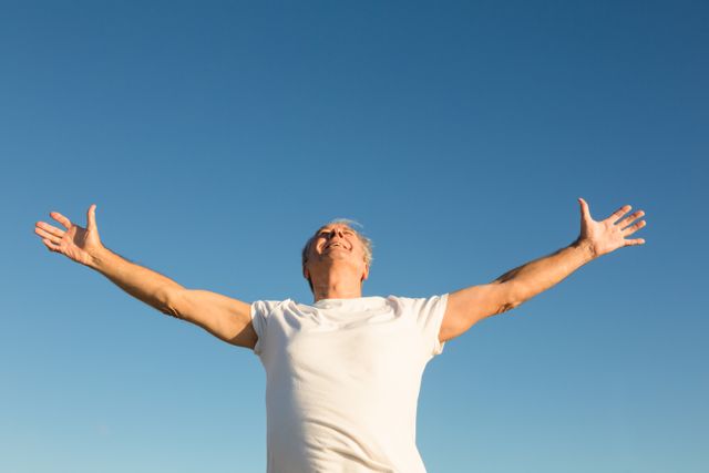 Senior man standing with arms outstretched under a clear blue sky, expressing freedom and joy. Ideal for use in advertisements promoting active lifestyles, wellness, retirement communities, or positive mental health. Can also be used in articles or blogs about aging gracefully, outdoor activities, and personal well-being.