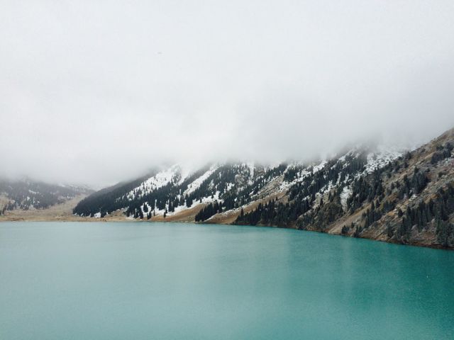 This image captures a tranquil mountain lake with turquoise waters, surrounded by snowy peaks partially shrouded in misty clouds. Ideal for travel brochures, nature blogs, and websites promoting outdoor adventure or tranquility.