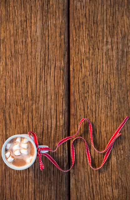 Cup of hot chocolate with marshmallows tied with a red ribbon on a wooden table. Perfect for holiday-themed promotions, Christmas cards, festive blog posts, and winter beverage advertisements.