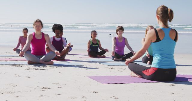 Group of women practicing yoga on beach, sitting in lotus position, meditating on sunny day with ocean in background. Perfect for promoting fitness retreats, outdoor wellness activities, healthy lifestyle.
