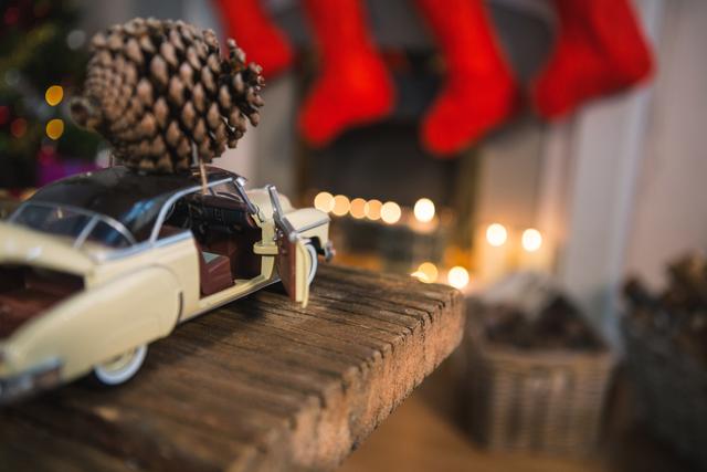 Toy car carrying pine cone placed on rustic wooden table in cozy Christmas setting. String lights and red stockings on fireplace in background create festive atmosphere. Ideal for holiday greeting cards, Christmas decoration inspiration, and festive home decor advertisements.