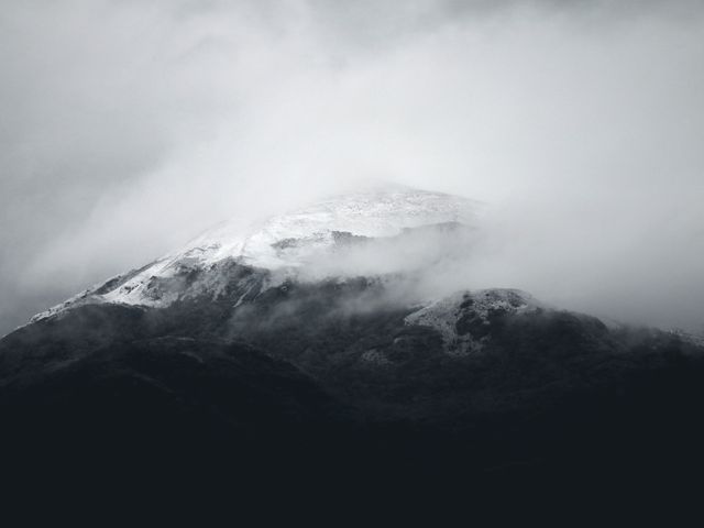 Black-and-white photograph of a majestic snow-covered mountain peak partially obscured by mist and clouds. The dark, rugged terrain contrasts with the white snow, creating a moody and serene atmosphere. Ideal for use in outdoor adventure promotions, travel imagery, posters, and nature-themed projects highlighting tranquility and remote wilderness.
