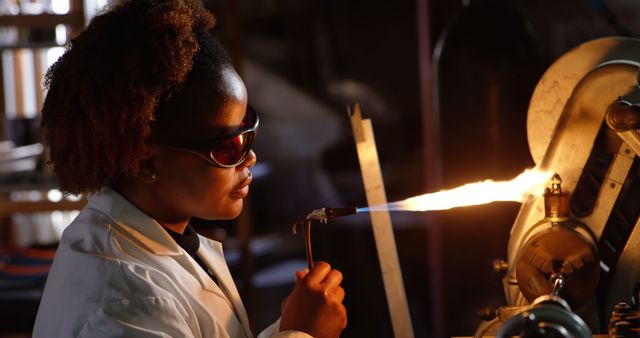 Female scientist works with a torch in a science lab for experiments or research. Wears a white coat and safety goggles for protection. Useful for illustrating scientific research, laboratory procedure, safety protocols, STEM education, or science-related articles.