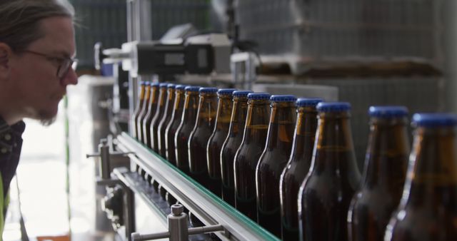 Caucasian male brewery worker wearing glasses looking at glass bottles in production line in brewery. Brewery, technology, production and food and drink, unaltered.