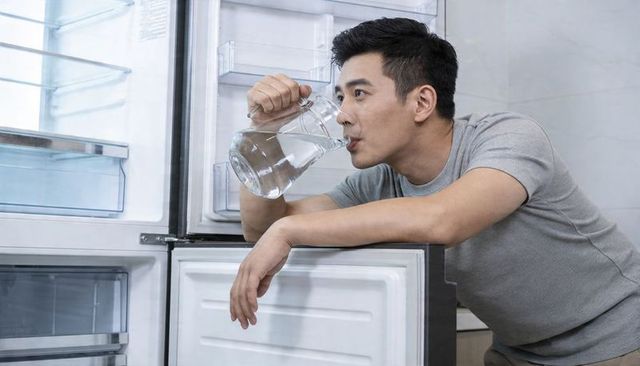 Man leaning on open refrigerator drinking water from glass pitcher in bright kitchen
