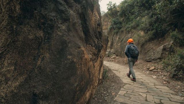 Solo hiker walking canyon trail by rugged rock face wearing orange cap and backpack