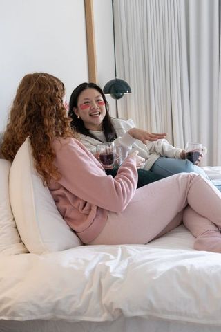 Cozy moment: diverse female friends reclining and chatting on bed holding clear glass mugs