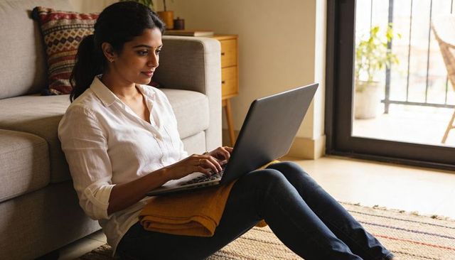 Young woman working remotely on laptop in cozy living room using mustard blanket