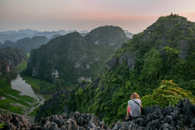 Woman sitting on rocky outcrop with lush green mountain range views during sunset. Ideal for travel blogs, adventure modelling portfolios, tourism advertisements, and outdoor lifestyle content.