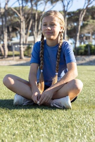 Preteen girl sitting cross-legged on sunlit grass wearing blue t-shirt and sneakers