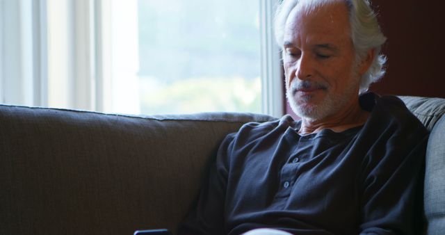 Senior man sitting comfortably on sofa, using smartphone, by bright window. Ideal for themes related to retirement, technology use among seniors, relaxing at home, and casual lifestyle photography.