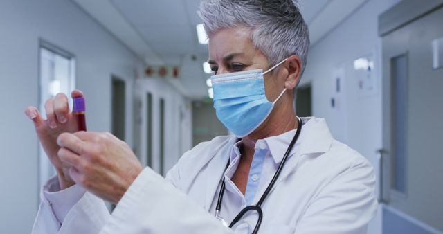 Senior female doctor wearing a surgical mask examining a blood sample in a hospital corridor. Ideal for illustrating themes related to healthcare, medical diagnosis, laboratory work, and professional medical services. Suitable for use in articles, websites, and brochures focused on health care and clinical environments.