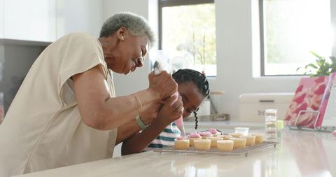 Senior african american grandma guiding granddaughter decorating cupcakes in modern kitchen
