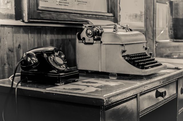 Black and white depiction of a vintage office showing a typewriter and a rotary phone on a wooden desk. This image is perfect for showcasing historical communication and typing technology, retro offices in films or advertisements, or evoking nostalgia in design projects.