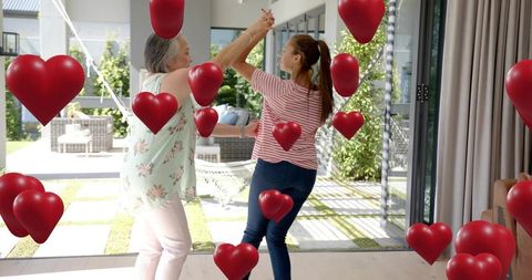 Mother and teen daughter dancing in sunlit living room with floating heart balloons