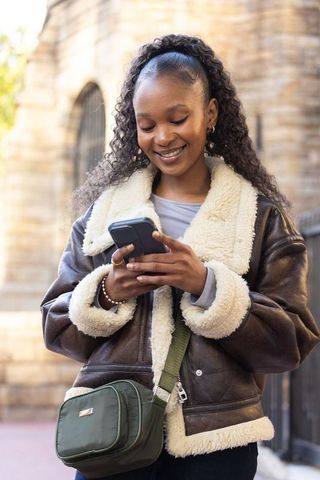 Young woman checking smartphone while smiling, wearing shearling jacket and green crossbody bag