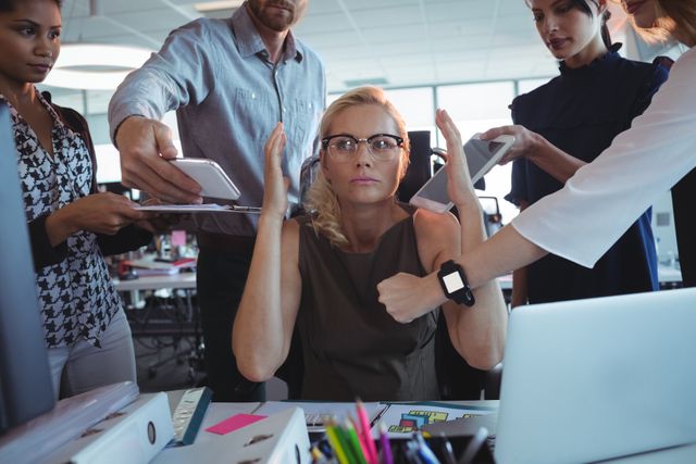 Businesswoman sitting at desk surrounded by colleagues holding various technologies, appearing stressed and overwhelmed. Ideal for illustrating workplace stress, multitasking challenges, and the pressures of modern corporate environments.