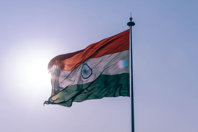 Indian flag waving in clear blue sky backlit by sun, evoking national pride and patriotism. Perfect for use in patriotic projects, cultural symbols representation, national day celebrations, and illustrating articles about India and its heritage.