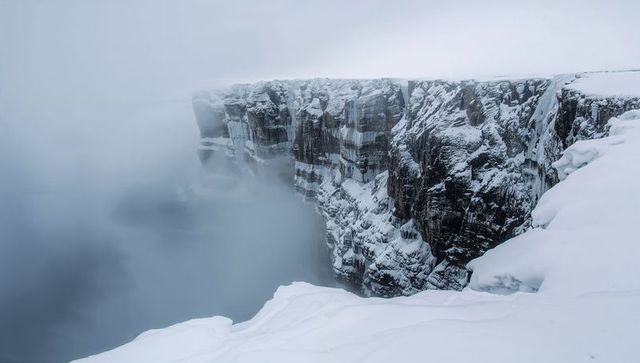 Snow-covered cliff rising above fog-filled gorge with hanging icicles and rock strata