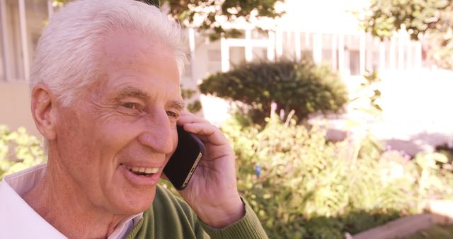 A senior man is smiling while talking on a phone in a garden with greenery in the background. Perfect for use in advertisements focused on elderly care, retirement plans, communication technology for seniors, and happy lifestyle depictions.